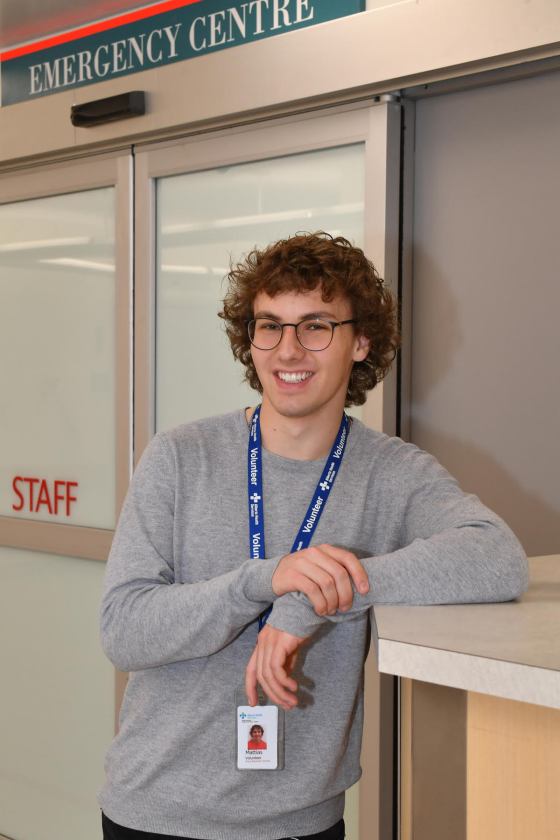 Smiling Mattias poses outside of an Emergency Centre entrance wearing an Alberta Help Services volunteer badge.